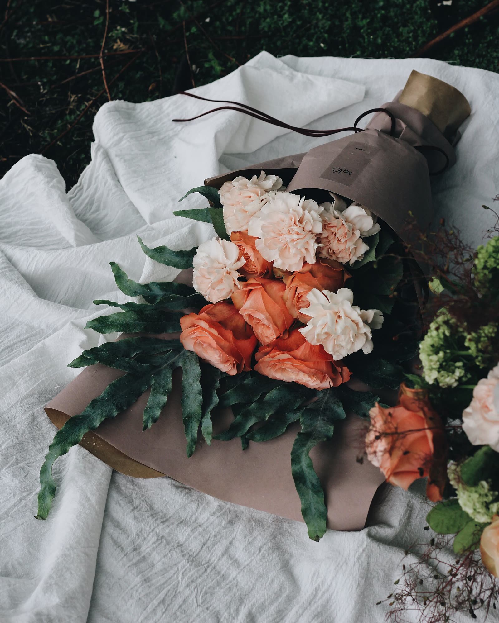 Buquê de rosas cor de pêssego e cravos brancos embrulhado em papel, deitado sobre um tecido de linho branco