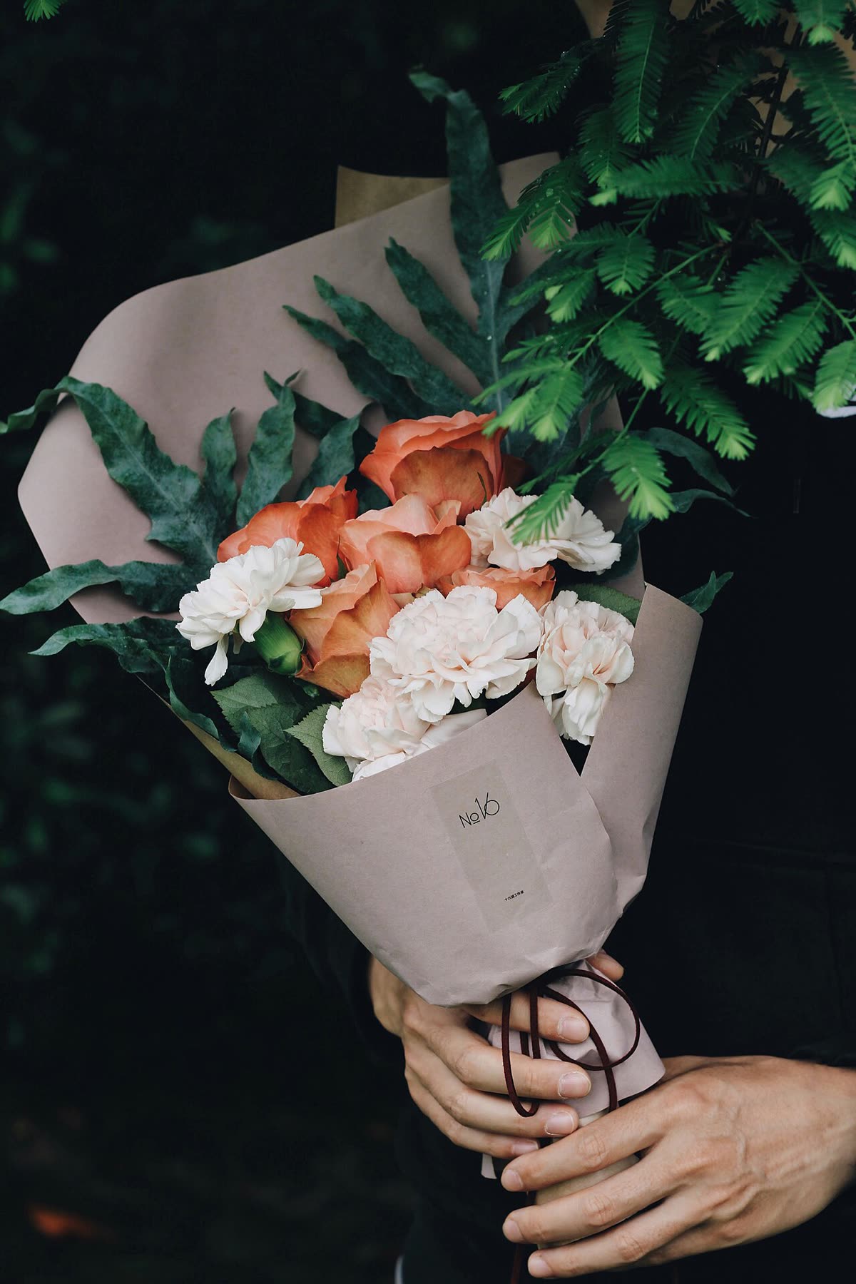 Mãos segurando um buquê de rosas e cravos embrulhado em papel rosado, com fita